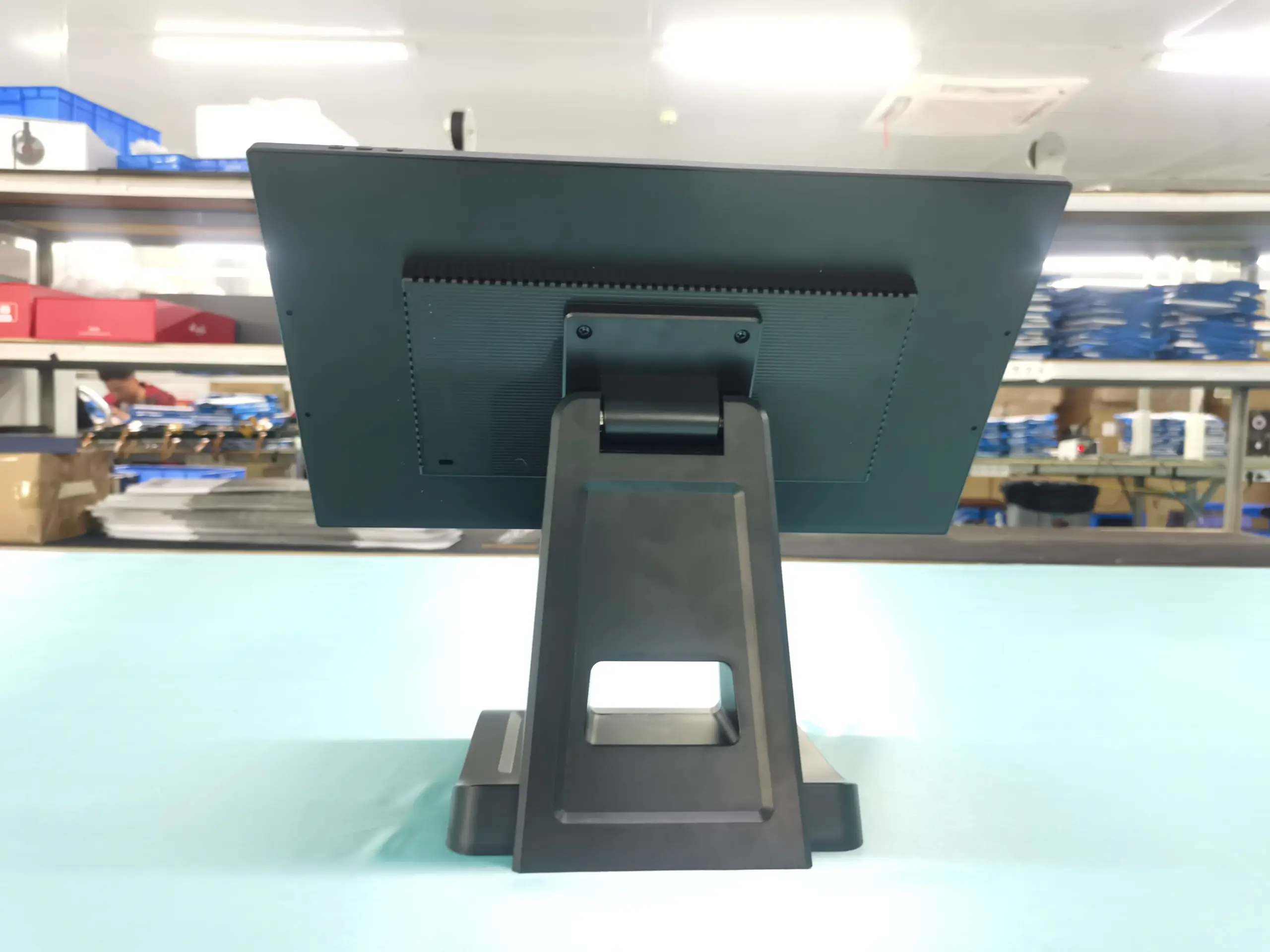 15.6 Inch All-in-One POS System Back View Rear view of a 15.6 inch black touchscreen POS system monitor on a sturdy stand in a factory setting.